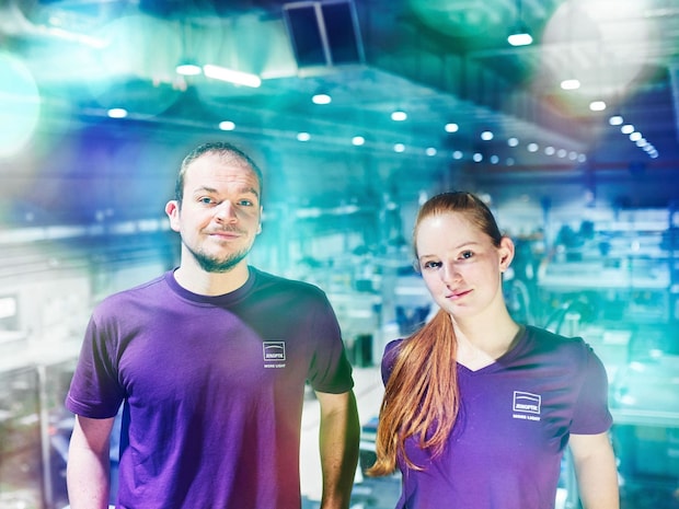 2 Jenoptik employees in violet shirt face the camera, production hall in the background