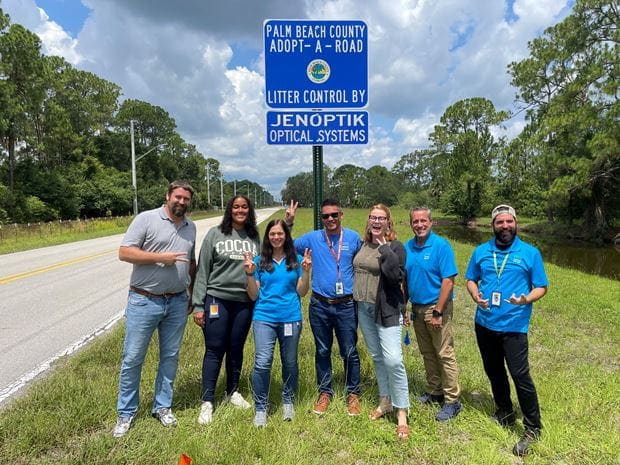 Jenoptik employees in front of a blue sign of the "Adopt a Road" program for litter control in Palm Beach County